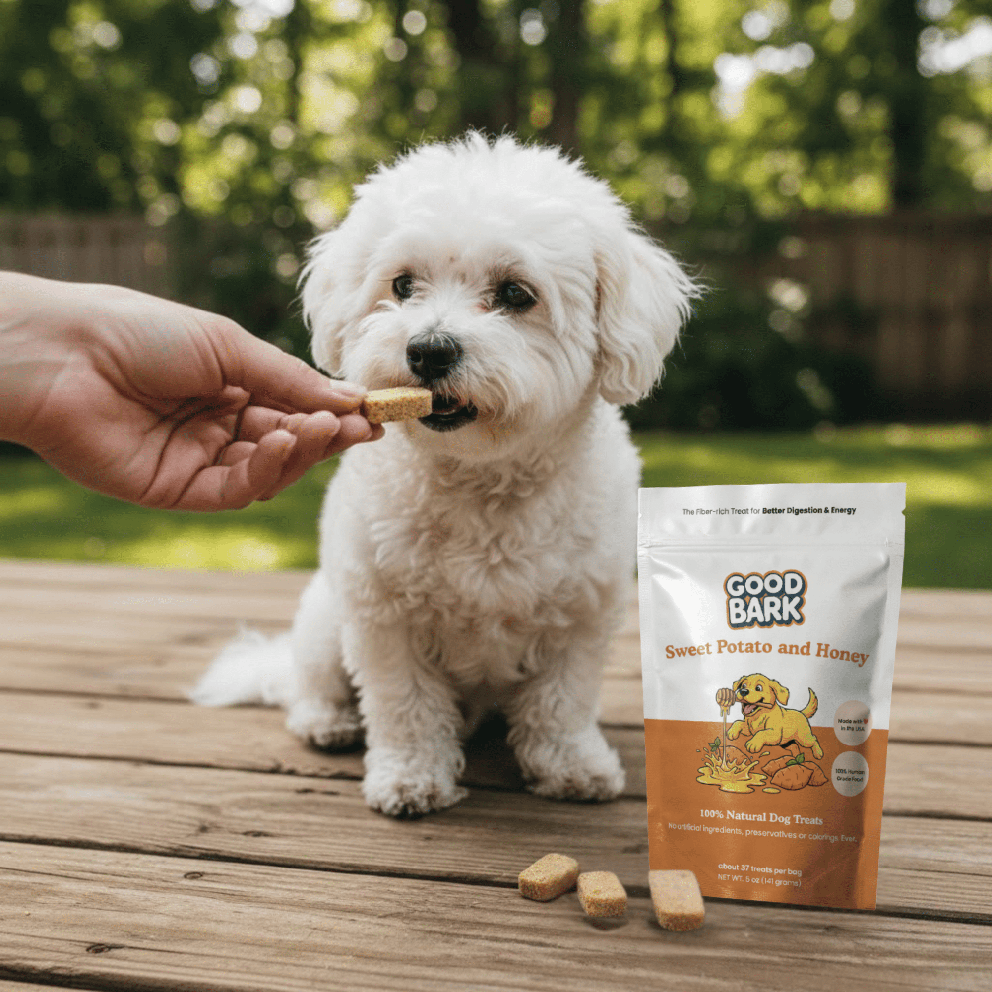 White dog being fed a treat by a hand, with a 'Good Bark' dog treat package on a wooden surface.